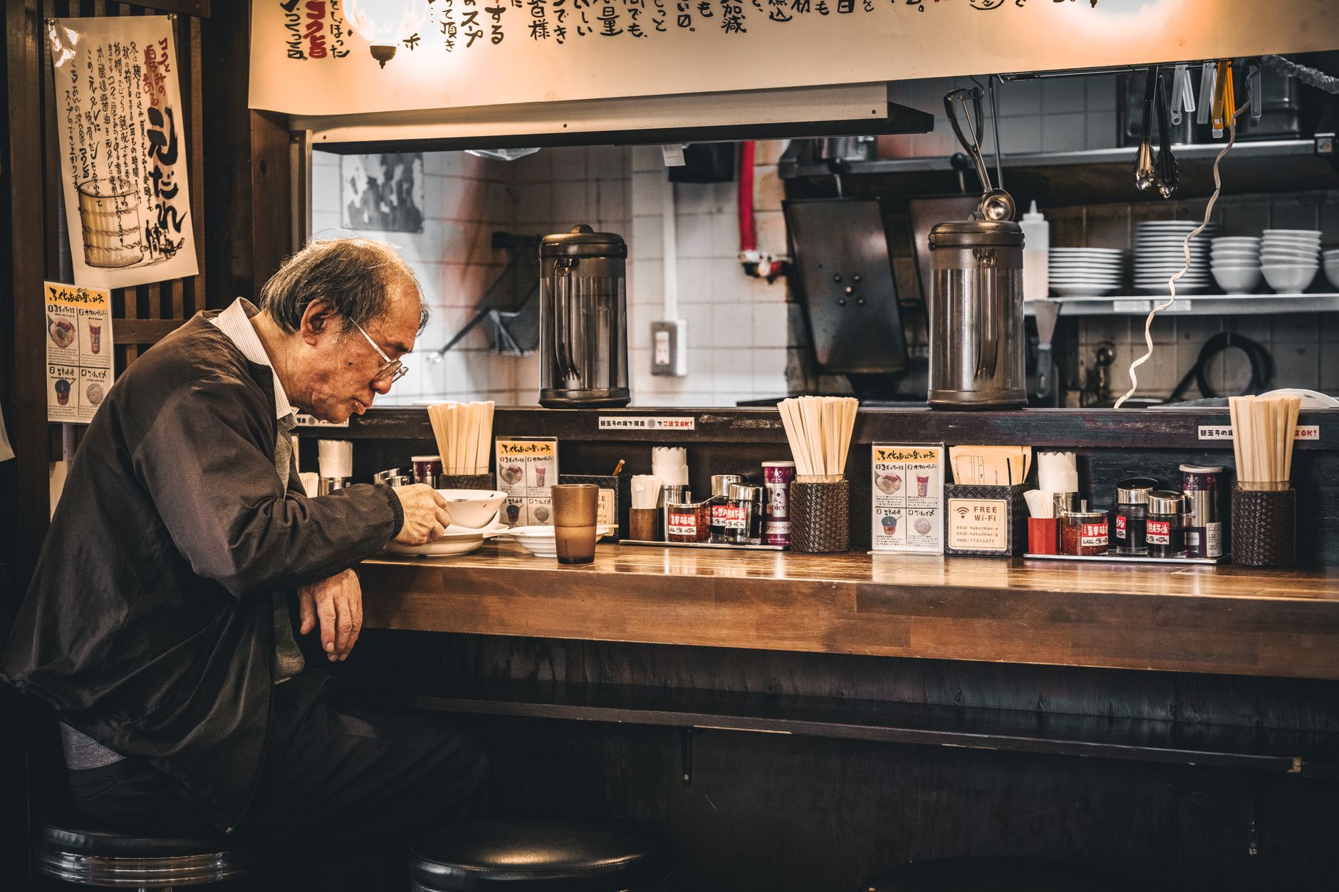A lively ramen shop counter with customers seated at the bar, steam rising from bowls of noodles in bright daylight