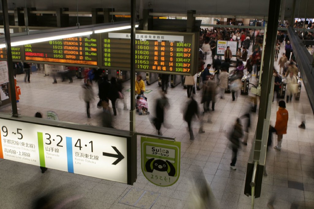 A busy Japanese train station concourse with platform and exit signage overhead.
